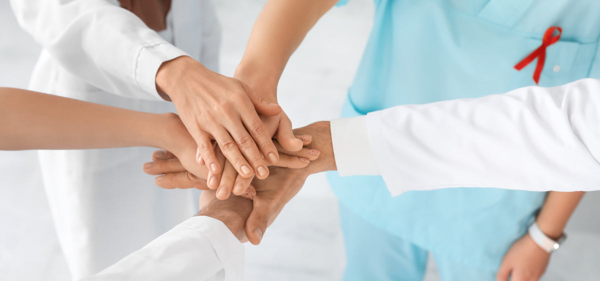 A group of healthcare workers stacking their hands together in a team gesture, with one person wearing a light blue scrub top and a red awareness ribbon pin, and another in a white lab coat.